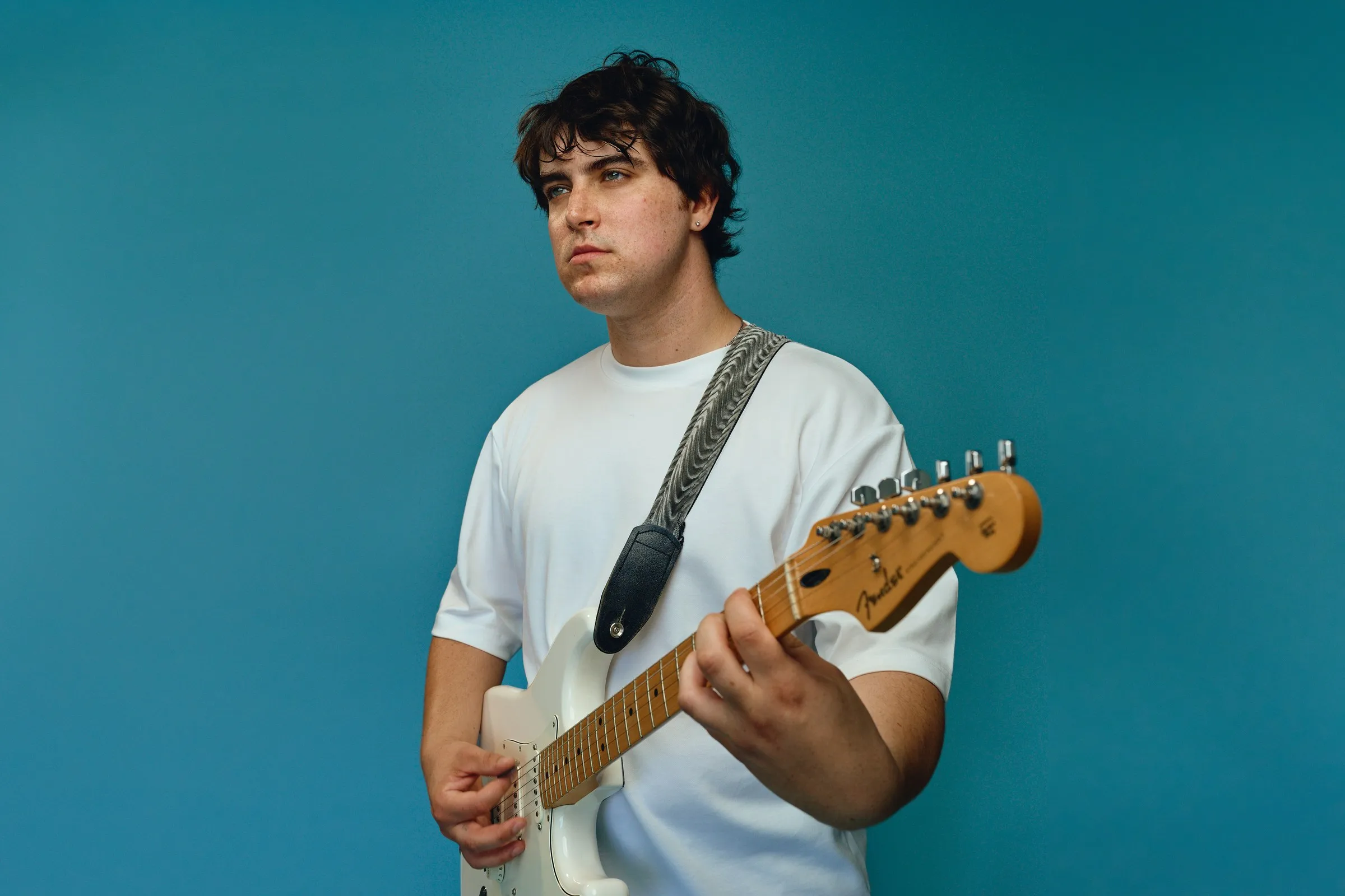 Sam Astra standing with a white Stratocaster in front of a teal backdrop