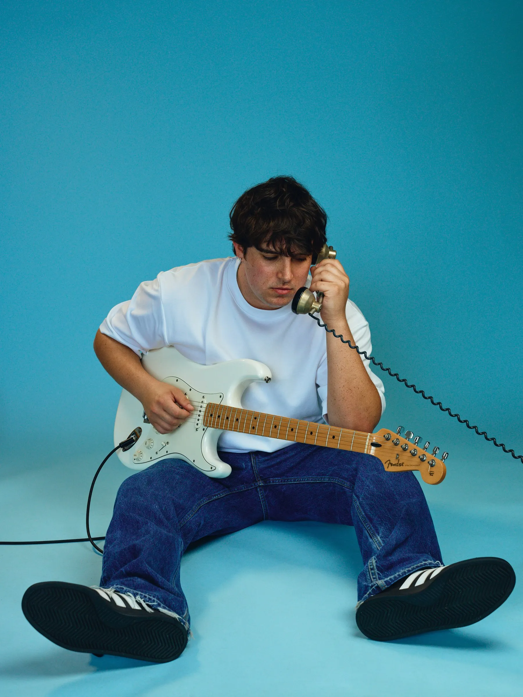 Sam Astra sitting on the floor with a white Stratocaster, holding a vintage phone to his ear, in front of a teal backdrop