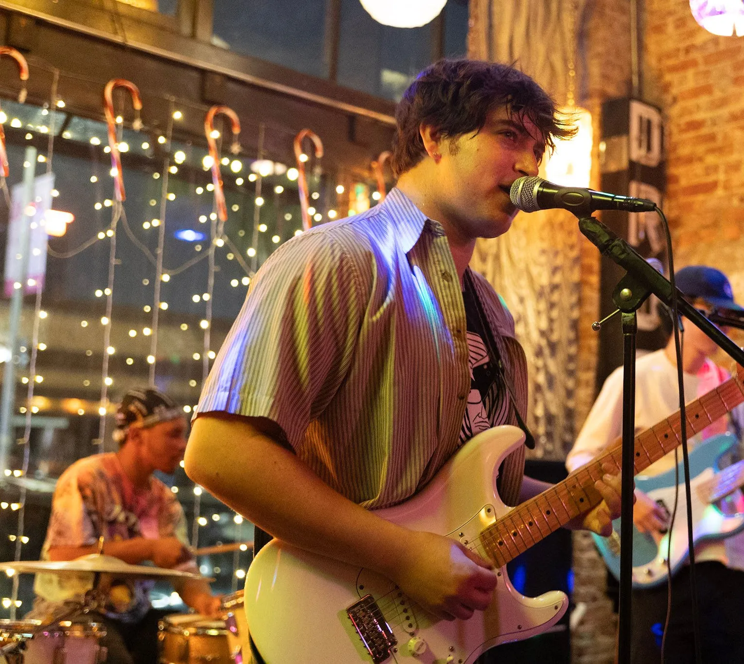 Sam Astra singing and playing a white guitar on stage in a bar lit with string lights, his band behind him