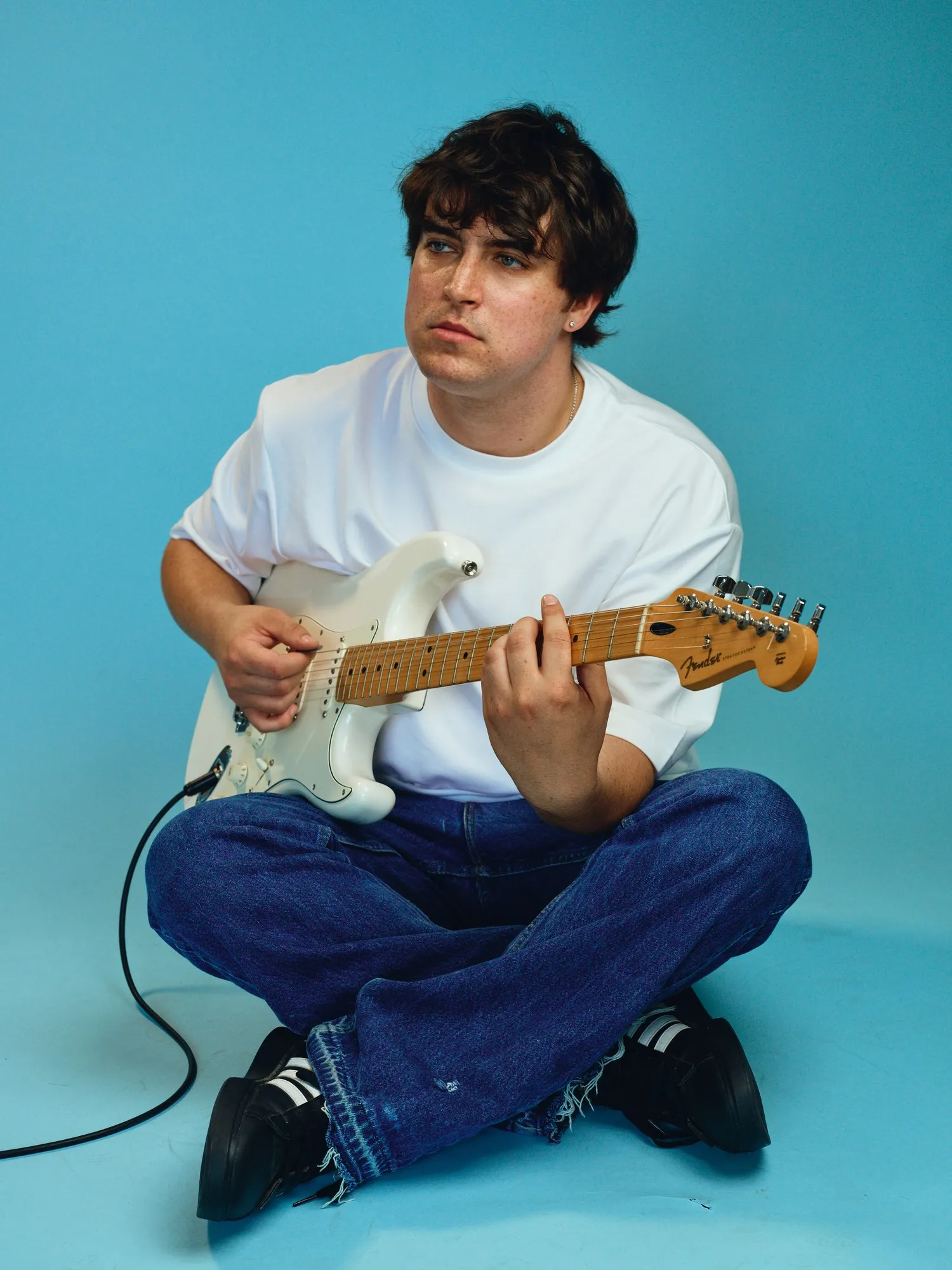 Sam Astra sitting cross-legged with a white Stratocaster in front of a teal backdrop